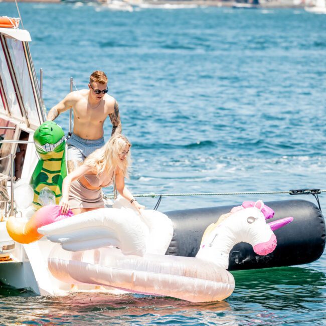 Two people descend from a luxury yacht hire in Sydney onto inflatable pool floats shaped like a unicorn and a llama, with a cityscape and sailboats in the background.