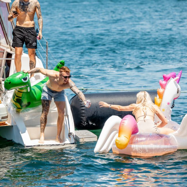 Two people enjoy a sunny day on inflatable floats next to a boat. One person on the boat helps another with an outstretched hand. A third person stands on the boat, highlighting the relaxed atmosphere of private yacht charter in Sydney Harbour.