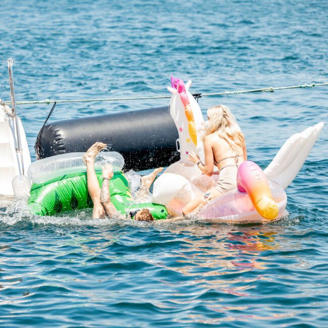 Several people are on inflatable floats next to a luxurious yacht in the water, with a person partially submerged, legs up, amid the floats. The scene appears lively and playful under a sunny sky – perfect for Corporate boat events Sydney or DJ boat hire Sydney.