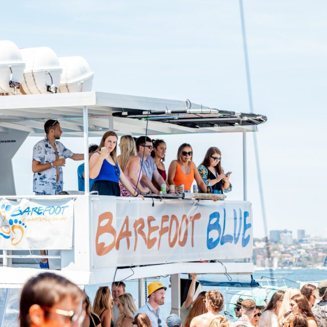 A group of people on the upper deck of a boat named "Barefoot Blue," watching an event on the water. Some are holding drinks, and others are leaning on the railings, enjoying what could be one of many corporate boat events Sydney offers.