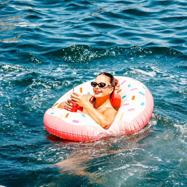 Person floating in water on a donut-shaped inflatable while holding a drink, wearing sunglasses, and smiling during a Sydney boat party hire.