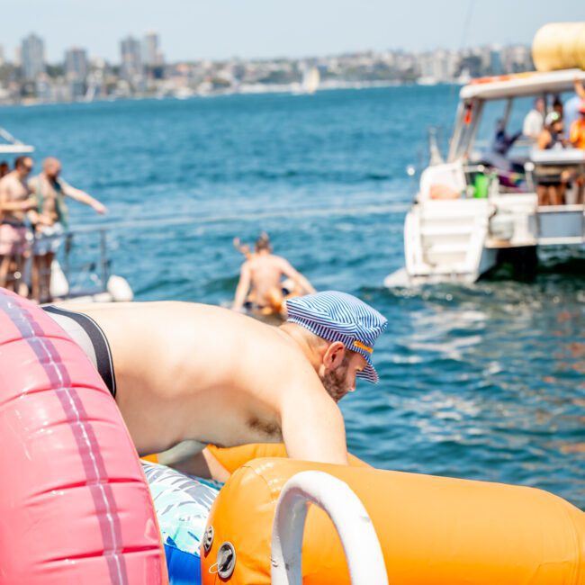 A man climbs onto an inflatable structure on water, surrounded by other people enjoying a DJ boat hire Sydney experience.