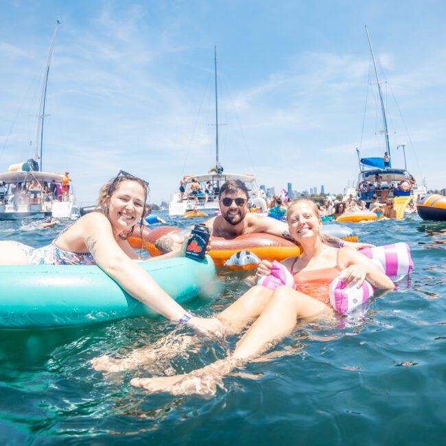 A group of people relax on inflatables in the water, surrounded by boats and other people, under a clear sky. A branded logo "The Yacht Social Club" is visible in the lower right corner, highlighting Luxury yacht hire Sydney.