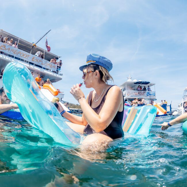 A woman in a black swimsuit and striped captain hat relaxes on an inflatable in the water, drinking from a cup. Several boats and people are visible in the background, enjoying what looks like a luxurious yacht hire Sydney experience.