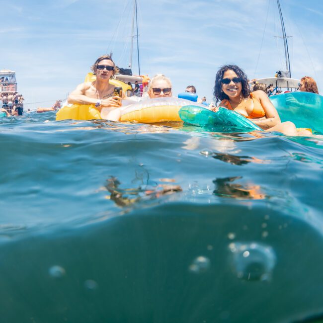 Three people on inflatable floats in the water, surrounded by boats and other individuals. The water level is up to the camera lens. A "Yacht Social Club" logo is visible in the bottom right corner, showcasing a luxurious Sydney boat party hire experience.