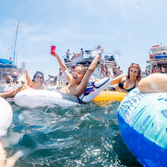 A group of people are floating on inflatable rafts and enjoying drinks in the water near several docked boats on a sunny day, reminiscent of a Sydney boat party hire.