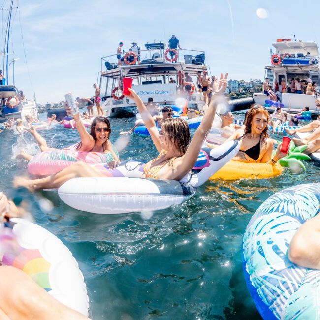 A group of people in swimwear are enjoying a sunny day in the water on inflatable floats, with several boats anchored nearby. They appear to be celebrating and having a good time, likely part of a luxury yacht hire Sydney experience.
