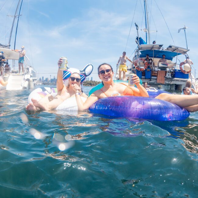 A group of people relax on inflatable floats in the water near anchored boats, enjoying drinks under a sunny sky during a luxury yacht hire in Sydney.