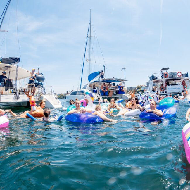 People relax on inflatable pool floats and interact in the water near several anchored boats on a sunny day, enjoying the perks of a private yacht charter around Sydney Harbour.