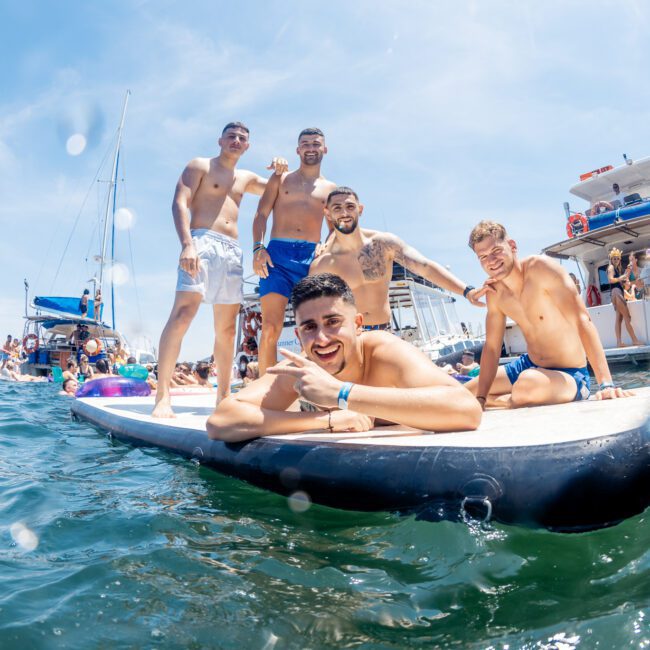 Five men are on a paddleboard in the water, posing for the camera with boats in the background. It is a sunny day. The Urban Social Club logo is present in the bottom right corner, highlighting their Sydney boat party hire service.