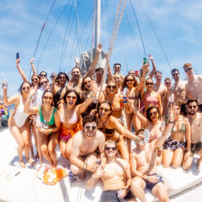 A large group of people in swimwear is posing and smiling on a private yacht charter in Sydney Harbour under a clear, sunny sky. They are holding drinks and making celebratory gestures. Other boats and water are seen in the background.