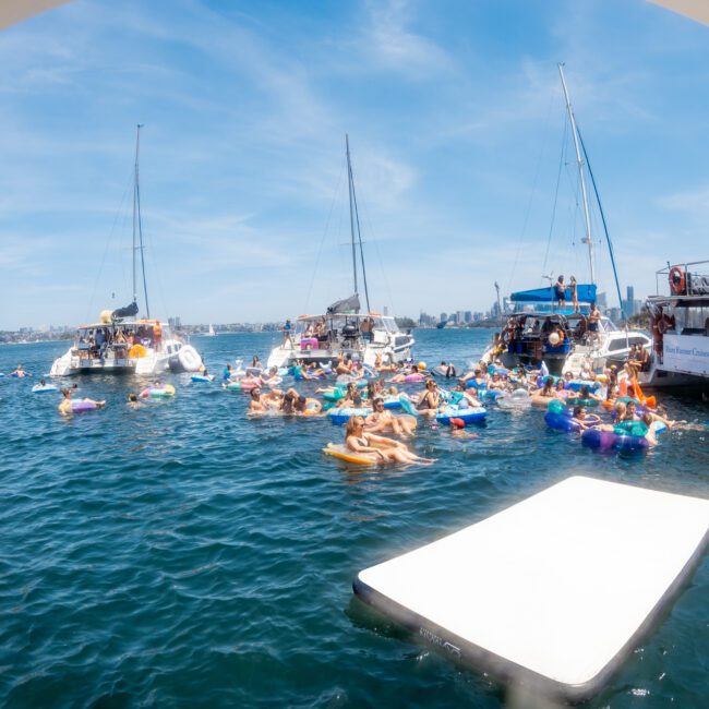 A group of people on inflatables gather between several anchored boats on a sunny day, enjoying a Sydney boat party hire.