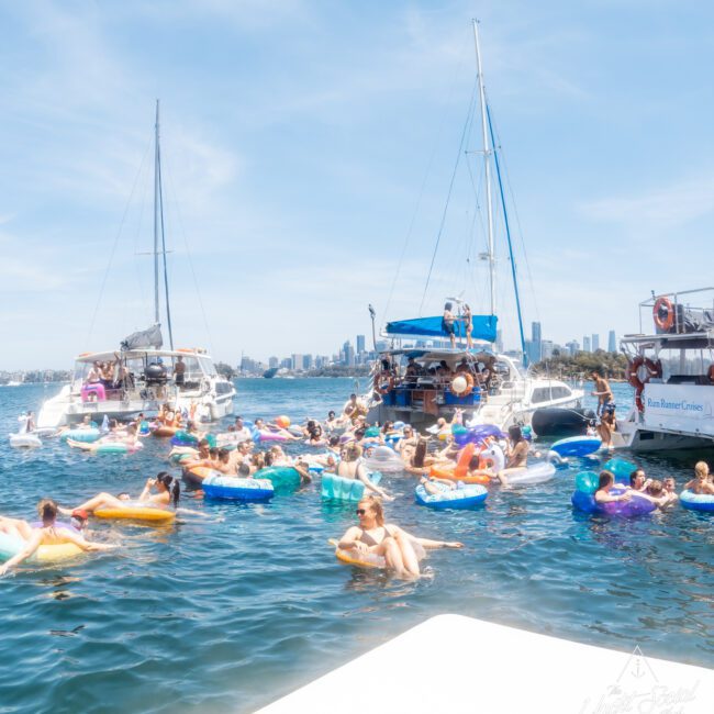 People are floating on inflatable tubes and swimming in a harbor near several boats on a sunny day. The city skyline is visible in the background, as a luxury yacht hire Sydney adds an elegant touch to the scene.