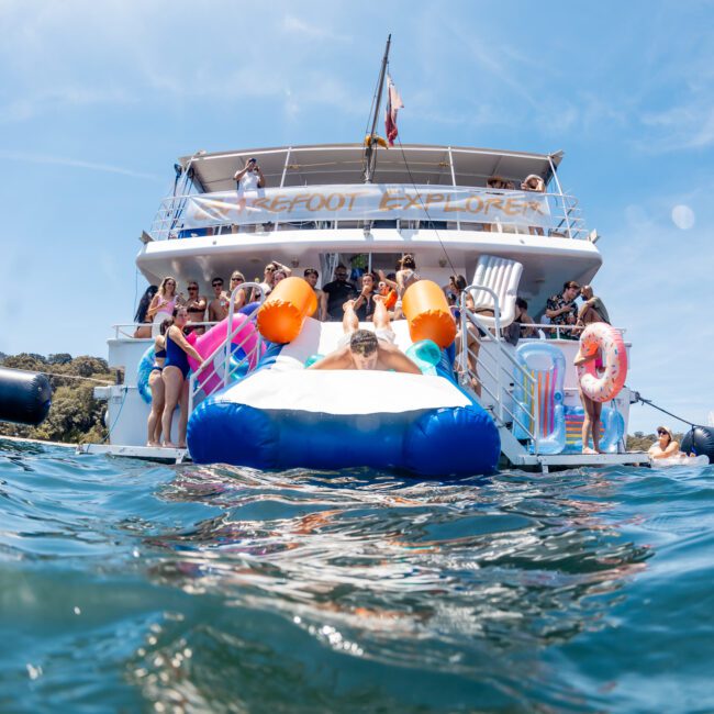 A group of people enjoys a sunny day on a boat with inflatable water toys, swimming in the sea. The image shows people lounging on the boat and floating near it during a Sydney boat party hire.