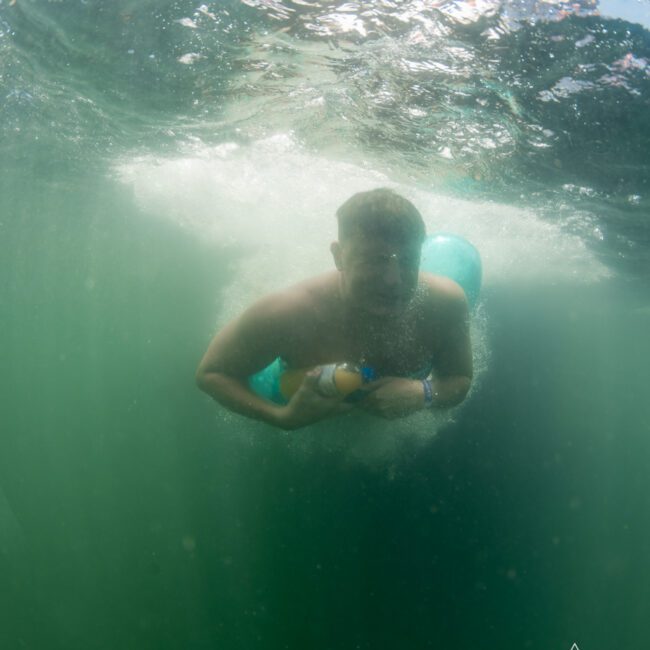 Person swimming underwater in a greenish body of water, holding a small object. Logo in the bottom right reads "The Yacht Social Club". Perfect for a Sydney boat party hire or DJ boat hire Sydney.