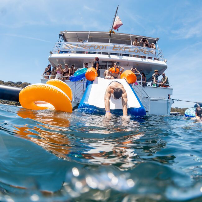 A group of people on a luxury yacht are enjoying a sunny day. Some individuals are using a water slide and inflatable tubes to glide into the water. The boat, named "Foot Explorer," is perfect for anyone looking for a Sydney boat party hire or private yacht charter in Sydney Harbour.
