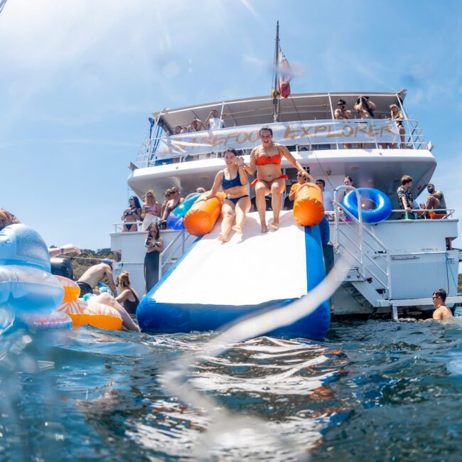 People on the "Explorer" slide into the water while others watch and swim nearby, enjoying a fun-filled Sydney boat party hire. Various flotation devices are visible in the sparkling blue water, enhancing the festive atmosphere.
