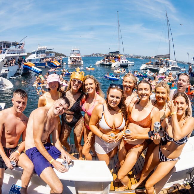 A group of people in swimwear pose for a photo on a boat. Several boats and people in the water are visible in the background under a clear blue sky, capturing the perfect moment at a Sydney boat party hire.