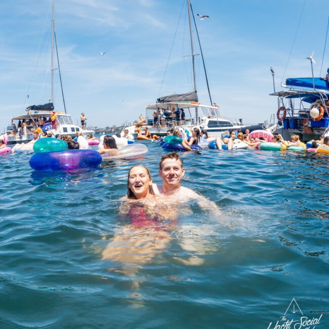 A man and woman smile while swimming in the water near a group of luxury yacht hires in Sydney. The scene is sunny with a clear blue sky, and other people are enjoying floats nearby.