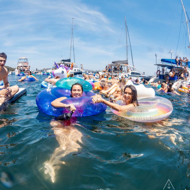 A group of people enjoy floating in a marina on inflatable pool toys, with sailboats and yachts anchored in the background. It's the perfect scene for a catamaran party in Sydney Harbour.