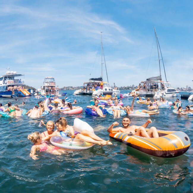 A group of people relaxing on inflatables and swimming in the water among several anchored boats on a sunny day, enjoying a vibrant catamaran party in Sydney.