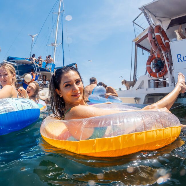 People floating on inflatable tubes in the water near anchored boats under a clear sky. One woman looks at the camera, while others enjoy the scene aboard the boats. "Rum Runner Cruise" is visible on one boat, making it feel like a private yacht charter Sydney Harbour event or even a DJ boat hire Sydney party.