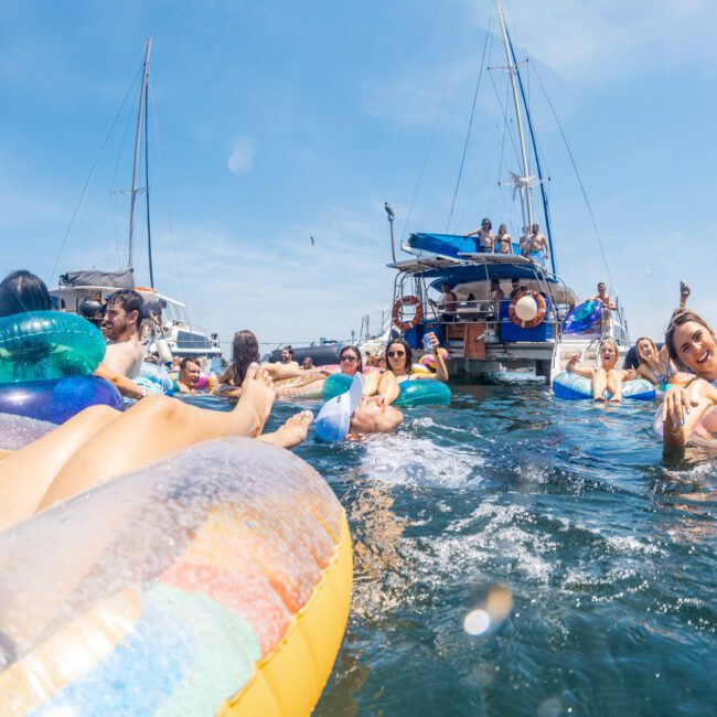 A group of people in swimsuits relaxing on inflatable tubes in the water near several boats on a sunny day, enjoying a catamaran party Sydney style.