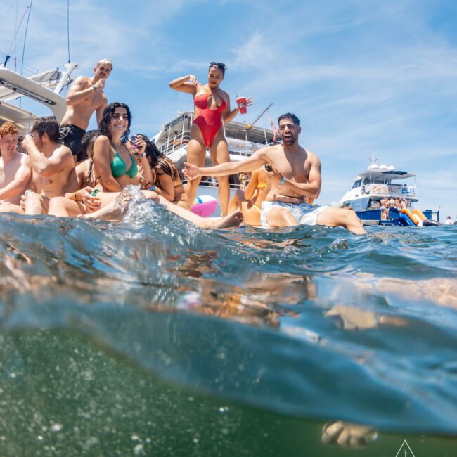 A group of people in swimwear is gathered on a private yacht charter on Sydney Harbour, enjoying drinks and dancing, with other boats visible in the background on a sunny day.