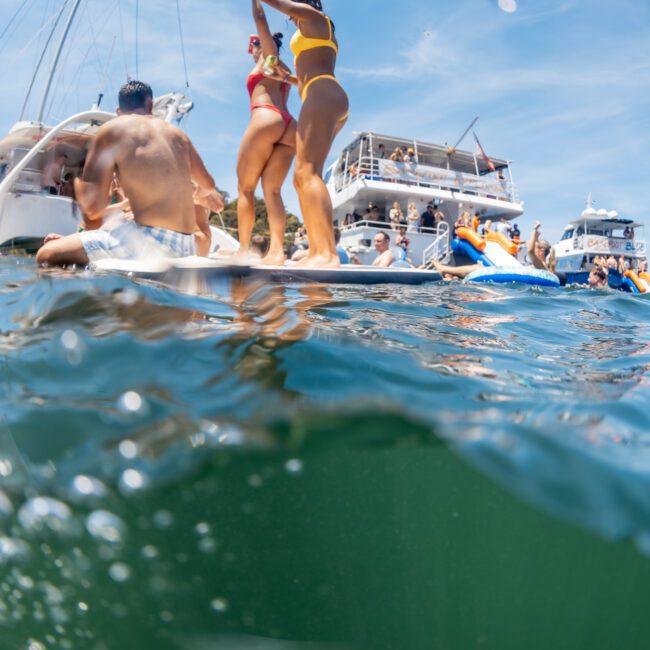Several people enjoy a day on the water, with two individuals in swimsuits on a boat and others nearby, surrounded by yachts. It looks like the perfect setting for a Sydney boat party hire or even corporate boat events in Sydney.
