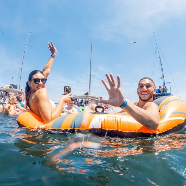 Two people in an inflatable ring wave to the camera while floating on the water. Several boats and other swimmers are visible in the background, perfect for a catamaran party Sydney experience.