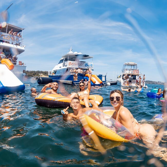 People are in the water and on inflatable floats near several docked boats with water slides on a sunny day. The atmosphere appears to be festive and relaxed, perfect for a luxury yacht hire Sydney experience or a private yacht charter Sydney Harbour adventure.