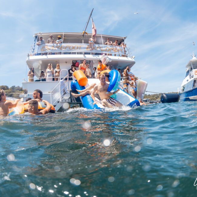 People are enjoying various water activities and inflatables near a large boat in a sunny outdoor setting, with onlookers on the boat and another vessel in the background, all part of an unforgettable Sydney boat party hire.