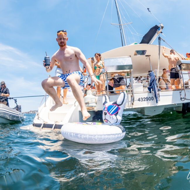 A man in swim trunks holds a drink while sitting on a white inflatable float shaped like a dog, next to a catamaran party in Sydney with people socializing on board.