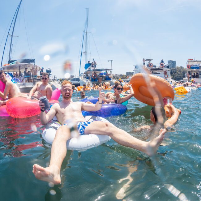 People are enjoying a sunny day on the water, lounging on inflatable tubes and floaties, with boats in the background. It's the perfect setting for a Sydney boat party hire or even a corporate boat event.