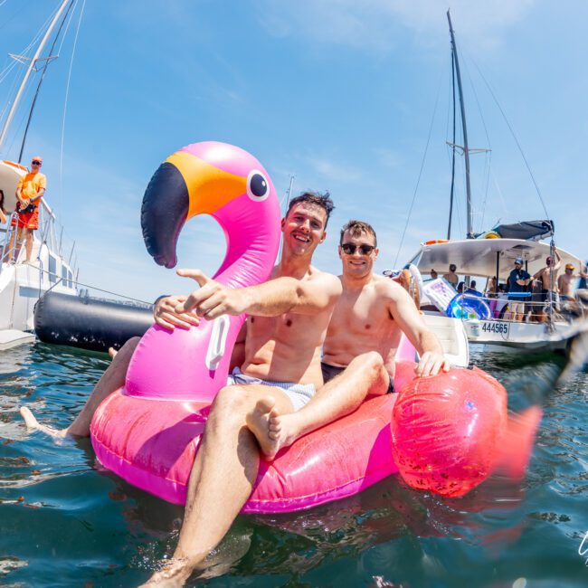 Two shirtless men smiling while sitting on a large pink flamingo float in the water, with boats and several people enjoying a Sydney boat party hire in the background.