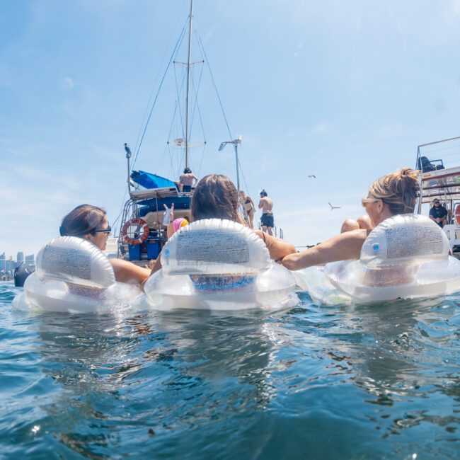Three people lying on inflatable floats hold hands in a body of water with boats and a city skyline in the background under a clear blue sky, enjoying a day that could be perfectly complemented by DJ boat hire Sydney.