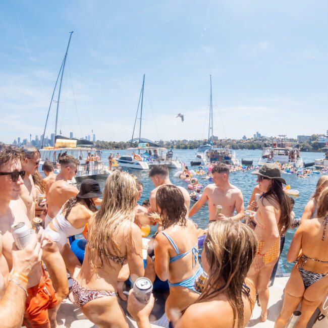 A group of people in swimsuits enjoy a boat party on the water, with yachts and cityscape visible in the background, exemplifying the fun and elegance of a private yacht charter on Sydney Harbour.