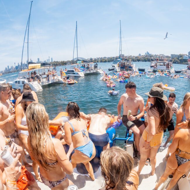A group of people in swimsuits are gathering on a boat, with additional boats and people floating in the water nearby. Some are holding drinks, and the skyline is visible in the background. This scene captures the essence of a vibrant Sydney boat party hire on a luxury yacht chartered around Sydney Harbour.