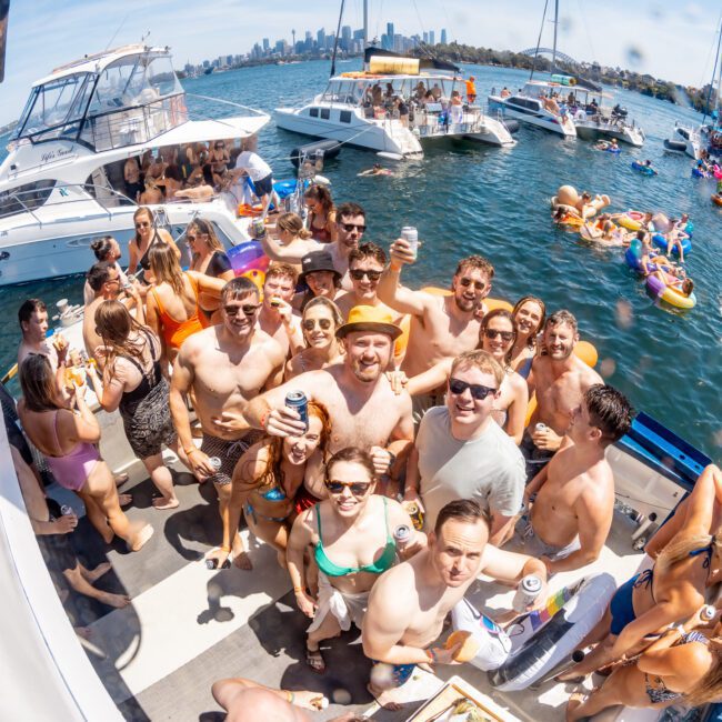 A group of people in swimsuits pose for a photo on a boat surrounded by other boats and people swimming in the water. The sunny sky and city skyline are visible in the background, perfect for a Catamaran party Sydney event or Private yacht charter Sydney Harbour.