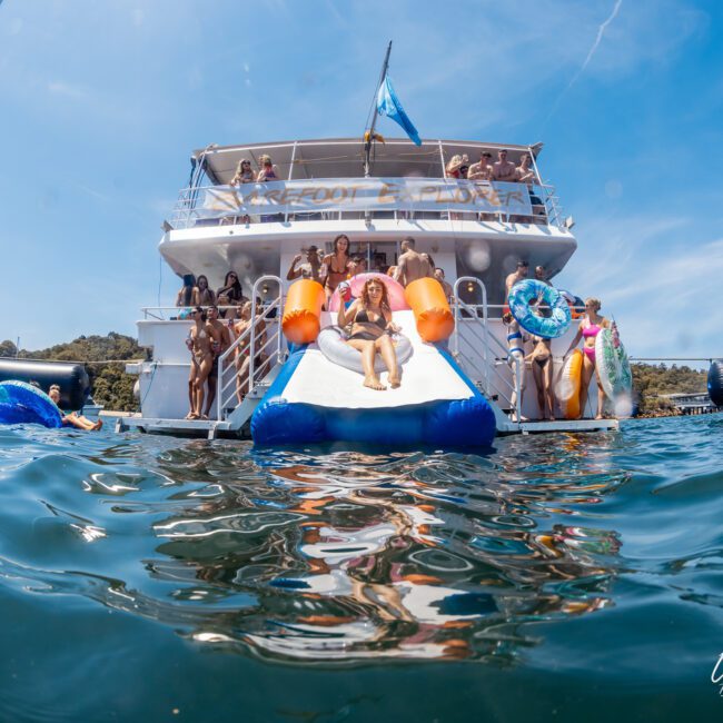 A group of people enjoys a sunny day on a boat. Some are on inflatables in the water, while others are on the boat's deck. There is a man sliding into the water from the back of the luxury yacht hire Sydney.