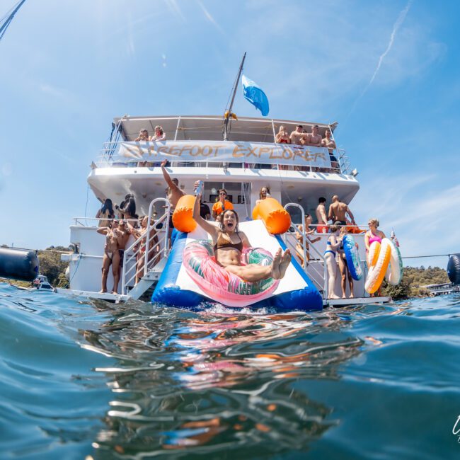 People enjoying a sunny day on a yacht, swimming and relaxing with floaties and inflatable toys. The luxurious yacht has multiple decks and a flag flying at the top. "The Yacht Social Club" logo is visible, perfect for Sydney boat party hire or corporate boat events Sydney.