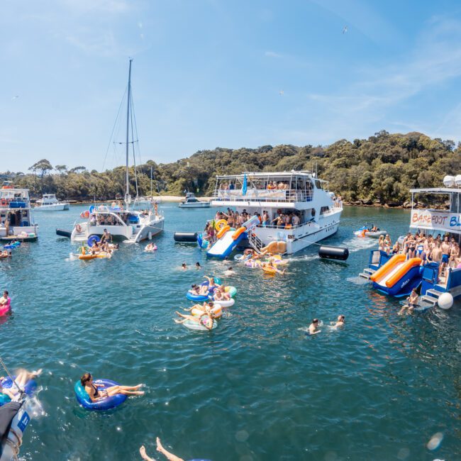 A group of boats are anchored close together in a bay with numerous people swimming, floating on inflatables, and using inflatable slides on a sunny day. Trees and a hill are visible in the background, creating an ideal setting for corporate boat events or private yacht charters in Sydney Harbour.