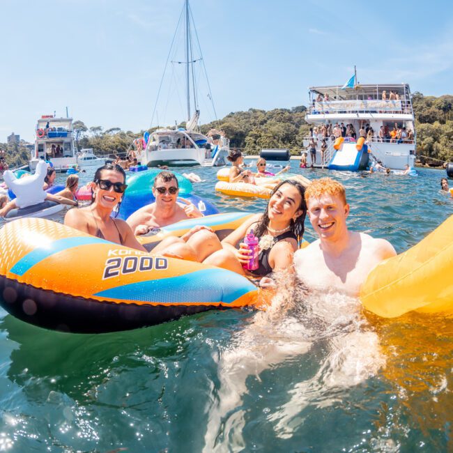 A group of people are floating on inflatable rafts together in the water near several anchored boats on a sunny day, enjoying a lively DJ boat hire Sydney event.