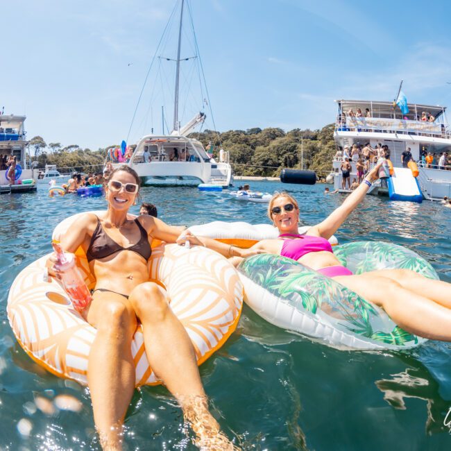 Two women relax on inflatable pool floats in the water, with boats and other people in the background. They are smiling and enjoying a sunny day, perhaps dreaming of a luxury yacht hire Sydney for an even more splendid experience.
