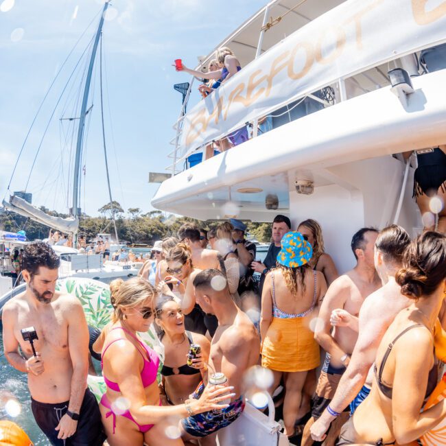 A group of people in swimwear are gathered and socializing on a catamaran under sunny skies. The boat is docked, and various individuals are chatting and holding drinks, enjoying the vibrant atmosphere of their Catamaran party Sydney.