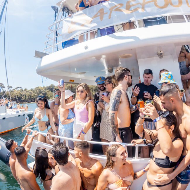 A lively group of people in swimsuits enjoy a catamaran party on Sydney Harbour with drinks in hand. Another boat is visible in the background on a sunny day, making it an ideal setting for corporate boat events.