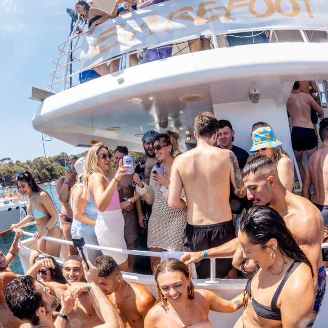 A large group of people partying on a yacht named "Barefoot," some in swimsuits, others holding drinks. The background shows calm waters and the shoreline, perfect for a catamaran party Sydney. The scene captures the vibrant atmosphere of a private yacht charter Sydney Harbour.
