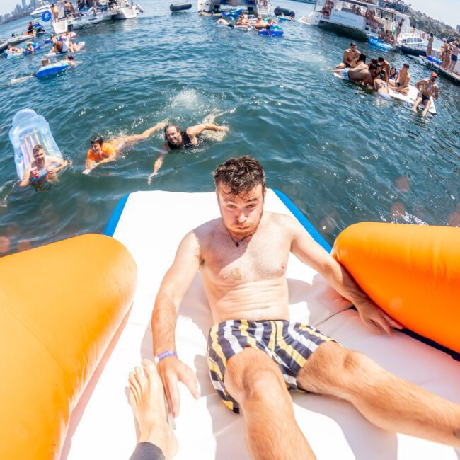 A man in swim shorts slides down an inflatable slide into a body of water surrounded by boats and people swimming and floating on inflatable devices, enjoying a vibrant catamaran party in Sydney.