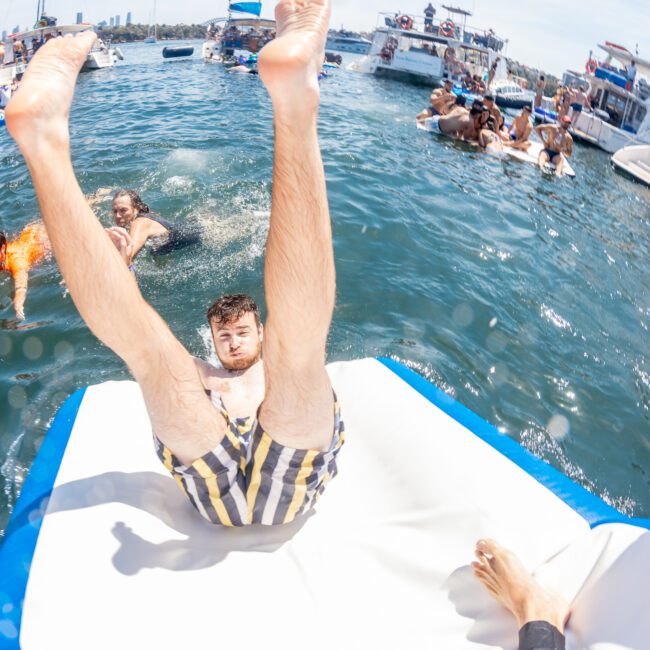 A man in striped swim trunks slides off an inflatable into the water, surrounded by other people swimming and boats in the background, epitomizing the fun of a Sydney boat party hire.
