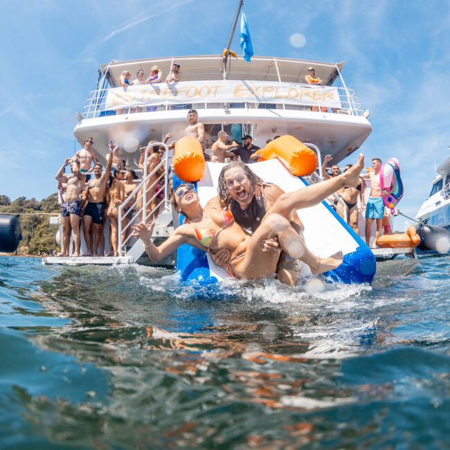 People sliding into the water from a luxury yacht named "AFLOAT EXPLORER" while others watch and take photos. Another luxury yacht with a similar name is seen in the background, showcasing the excitement of a private yacht charter on Sydney Harbour.
